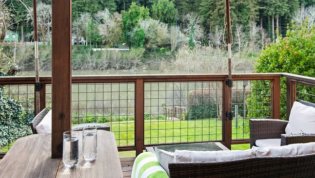 A wooden deck with a table and chairs, a red umbrella, and a view of a grassy area with trees. - River Time ~ Come Stay on the River! Access to Park, Riverfront, In Town (Monte Rio)