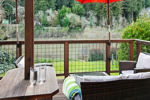Una terraza de madera con una mesa y sillas, un paraguas rojo y vistas a un área verde con árboles.