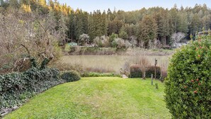 A serene lakeside garden with a grassy area, a small wooden bridge, and dense forest in the background. - River Time ~ Come Stay on the River! Access to Park, Riverfront, In Town (Monte Rio)