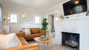 A living room with a white fireplace, a brown leather sofa, a round mirror, and a television mounted on the wall. - Cozy Cottage across from Grand Haven Beach (Grand Haven)