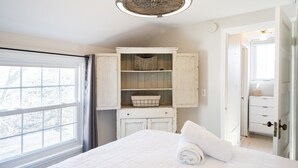 A bedroom with a large window, a white bed with a rolled towel, a wooden cabinet with shelves, and a door leading to another room. - Cozy Cottage across from Grand Haven Beach (Grand Haven)