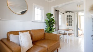 A living room with a brown leather sofa, a round mirror, a large green plant, and a white dining table with two chairs. - Cozy Cottage across from Grand Haven Beach (Grand Haven)