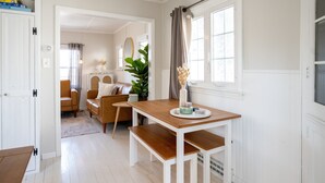 A dining area with a wooden table and benches, a white door, and a view into another room with a sofa and a plant. - Cozy Cottage across from Grand Haven Beach (Grand Haven)