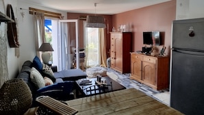 A cozy living room with a sofa, coffee table, and a TV. There's a kitchen area with a fridge, cabinets, and a dining table. - Maison Entre LAC ET Ocean A Hourtin EN Gironde, Idéal Avec de Jeunes Enfants ! (Hourtin)