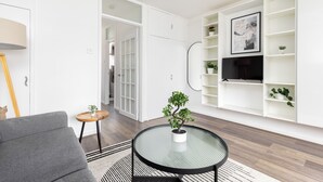 A modern living room with a grey sofa, a round glass table with a potted plant, and a white entertainment unit with shelves and a TV.