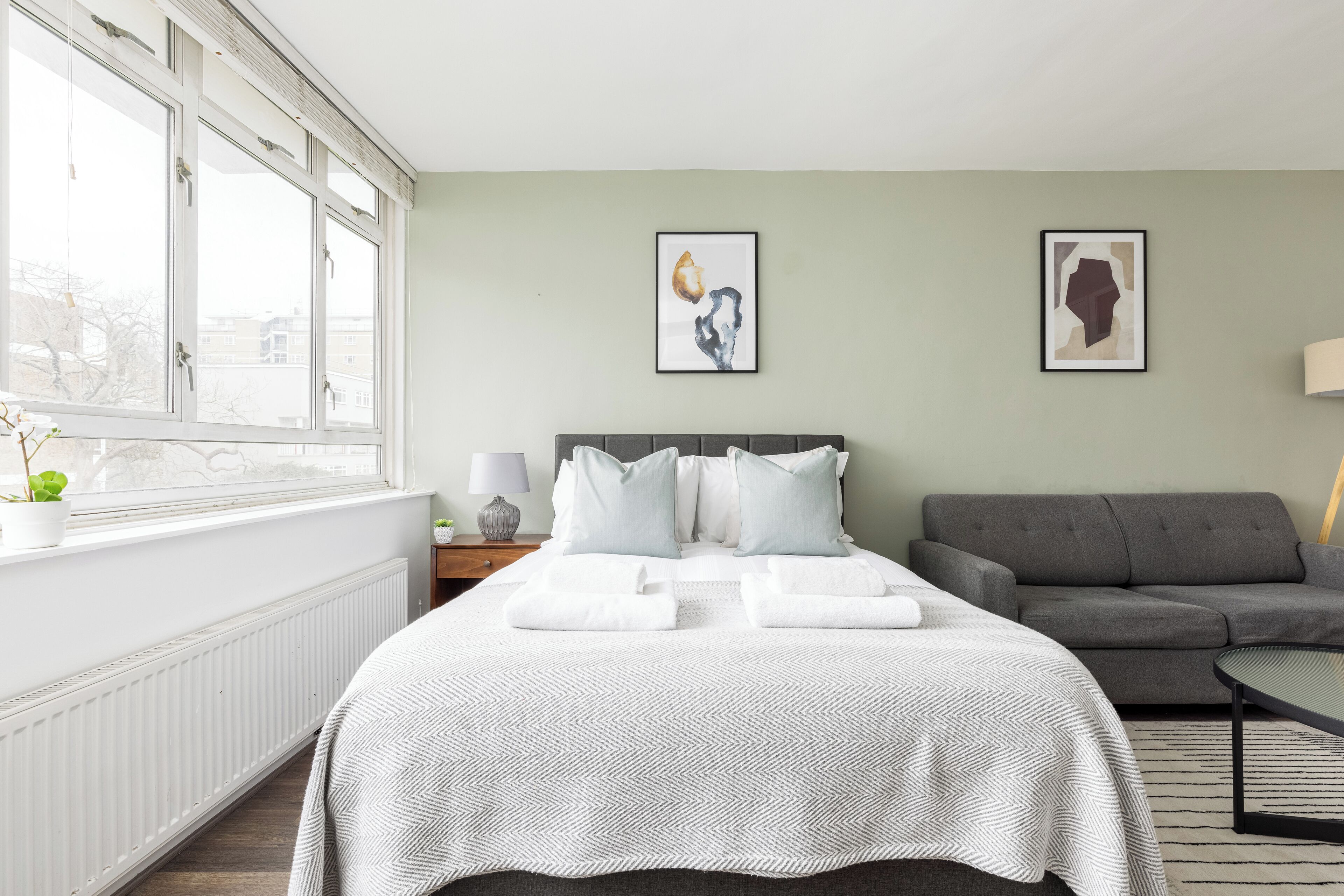 A hotel room with a large bed, a grey sofa, two framed artworks on the wall, and a window with a view of buildings.