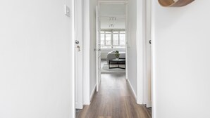 A hallway with white walls, a wooden floor, and a view into a bedroom with a bed, a small table, and a window.