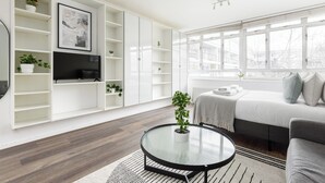 A modern living room with a glass coffee table, a sofa, a TV on a white cabinet, and a large window with a view of buildings.