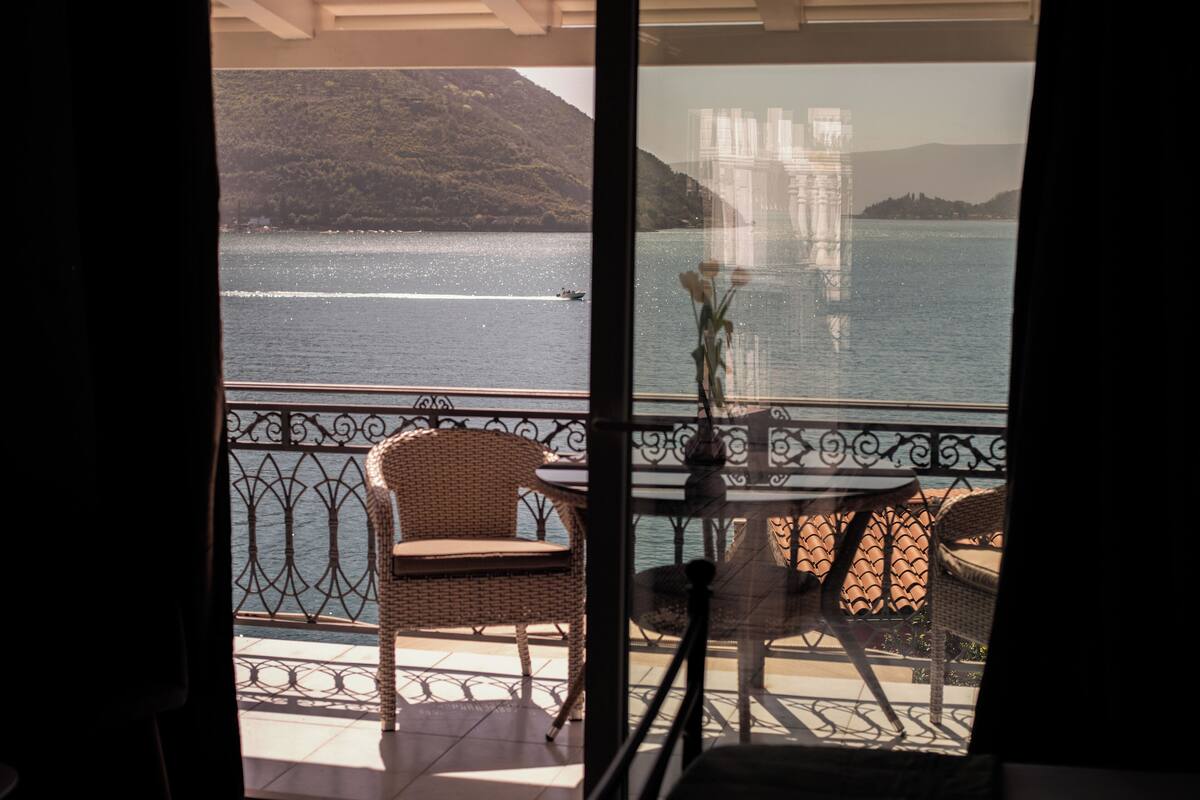 En balkon med fletstole og et bord, med udsigt over vand med både og fjerne bakker.