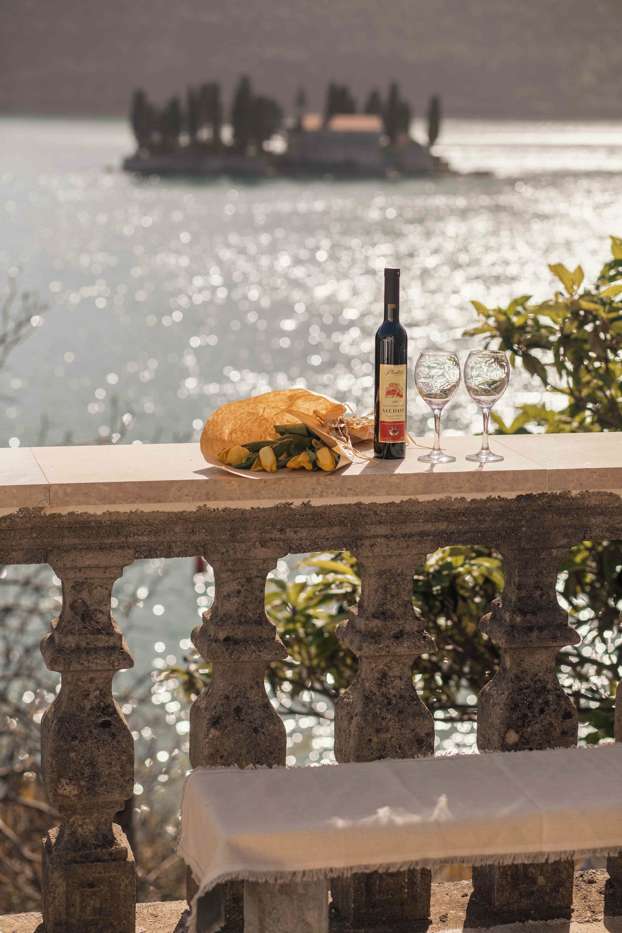 A table with a bottle of wine, two glasses, and a loaf of bread on a balcony overlooking a body of water.