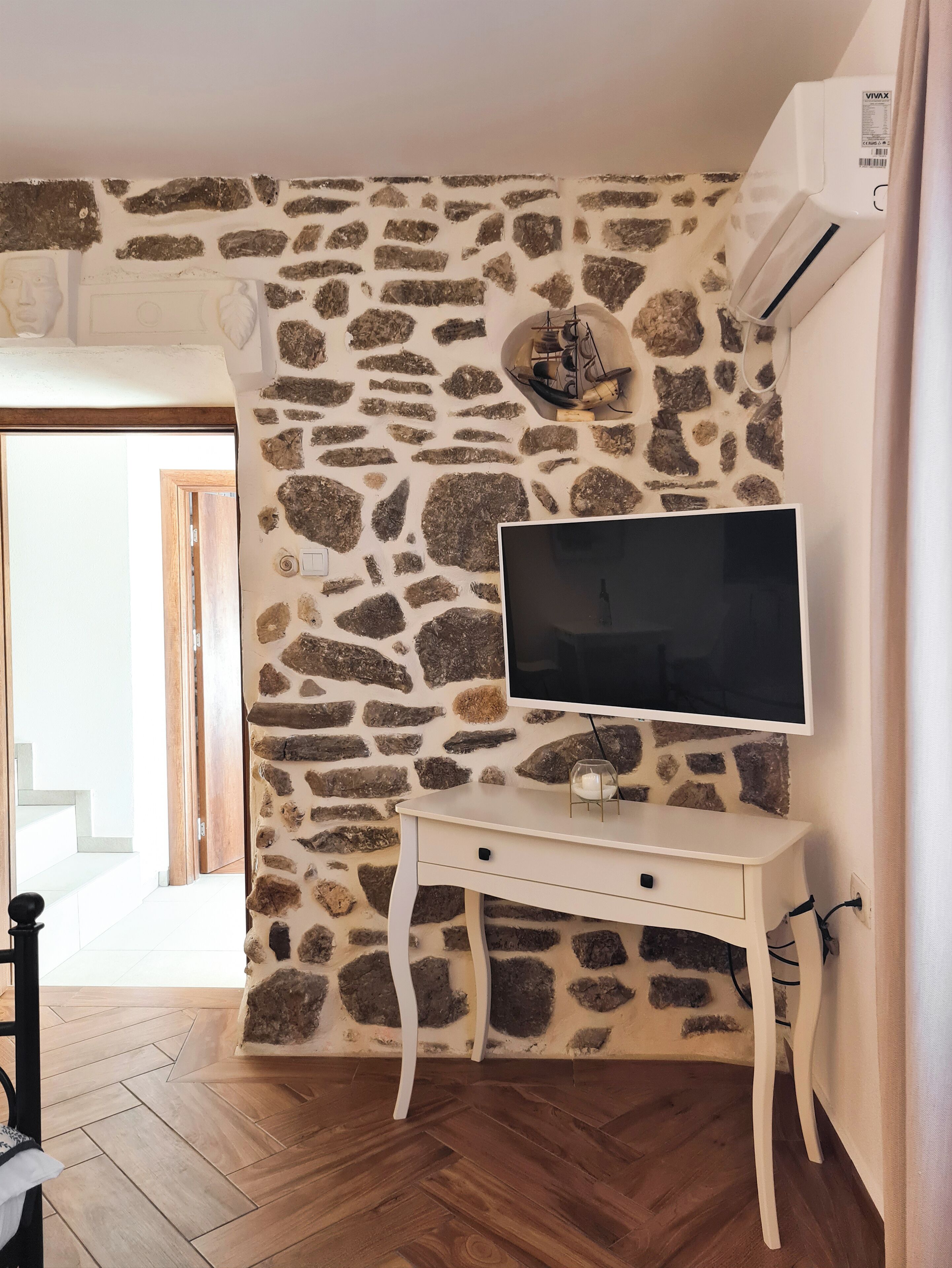 Quarto com parede de pedra, televisão de tela plana sobre um console branco e piso de madeira.