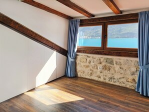A room with a stone wall, wooden beams, a large window with curtains, and a view of a body of water.