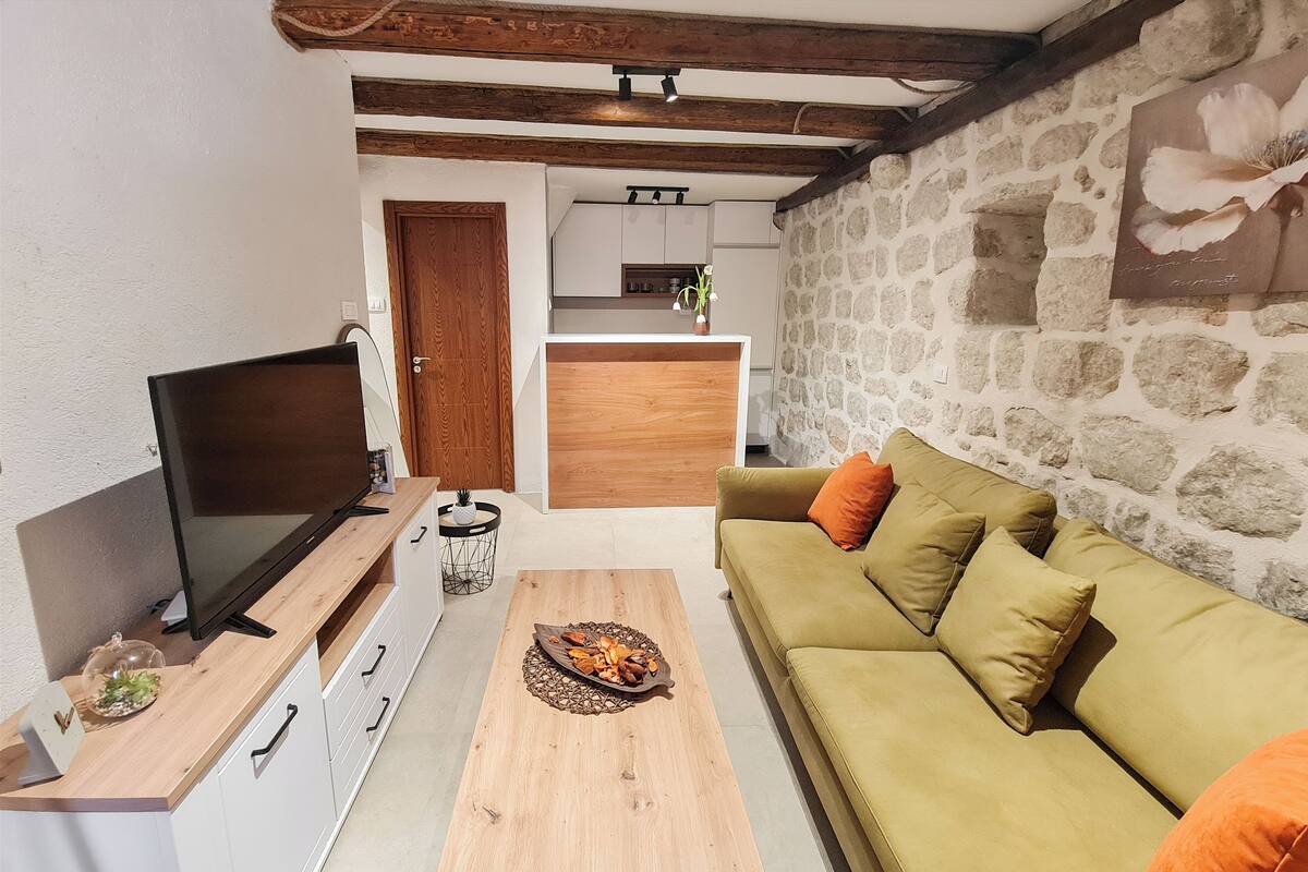 En hyggelig stue med et fjernsyn, et træsofa og en sofa med orange puder. Et køkken kan ses i baggrunden.
