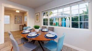 A dining area with a wooden table set for four, surrounded by chairs, a potted plant, and a window with a view of a house and palm trees. - Carlsbad Beach Bungalows - 4BRs, Outdoor Living, Walk to Beach, Pet-Friendly (Carlsbad)