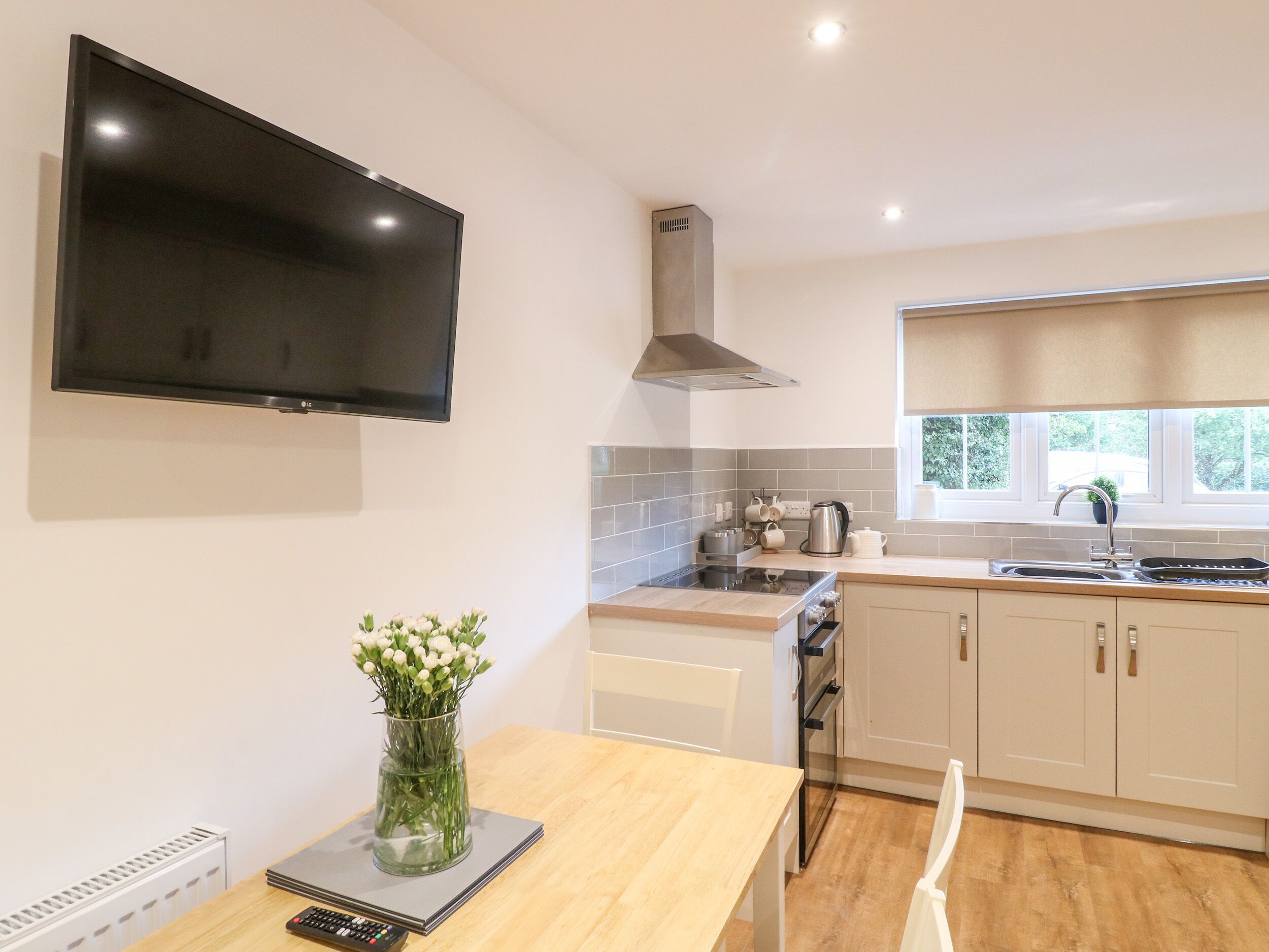 A modern kitchen with a flat-screen TV, a stainless steel range hood, a window with a view, and a vase with flowers on the counter.