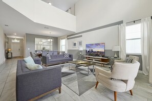 A spacious living room with a grey sofa, a coffee table, and a TV. There is a dining area with a table and chairs, and a kitchen in the background. - 5 Bed In Champions Gate, Modern Decor, Kid Themed Rooms, Game Room, Gated Resort! 5 Bedroom Home by RedAwning (Davenport)