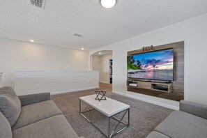 Uma sala de estar moderna com televisão de tela plana fixada em uma parede de madeira, uma mesa de centro branca e sofás cinzas.