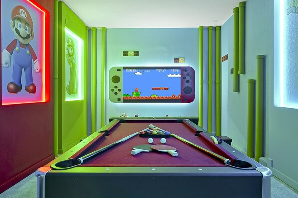 Uma mesa de bilhar com uma tela de videogame ao fundo, cercada por paredes verdes e uma decoração temática do Mario.