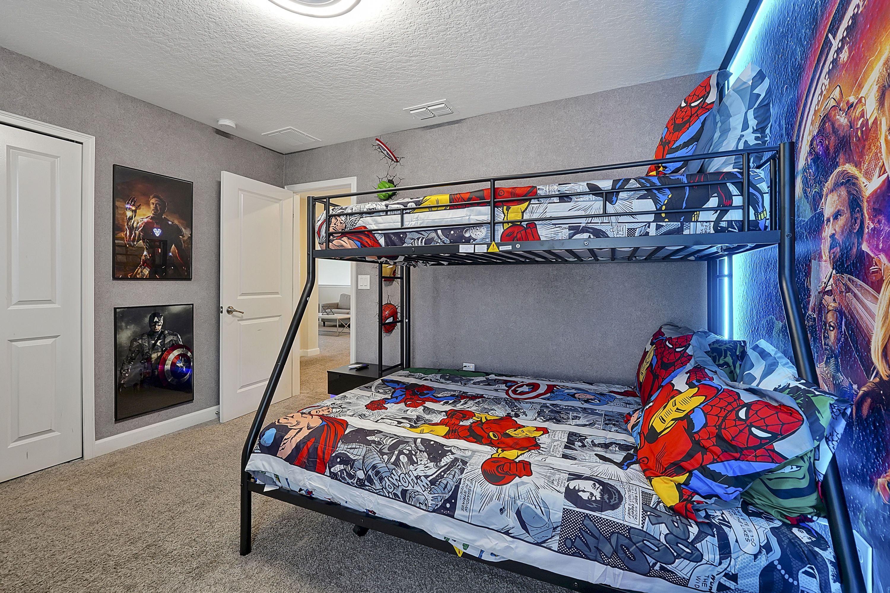 Un lit superposé avec une housse de matelas aux couleurs de Spider-Man, un mur orné d’une affiche de super-héros et une porte avec un tableau encadré.