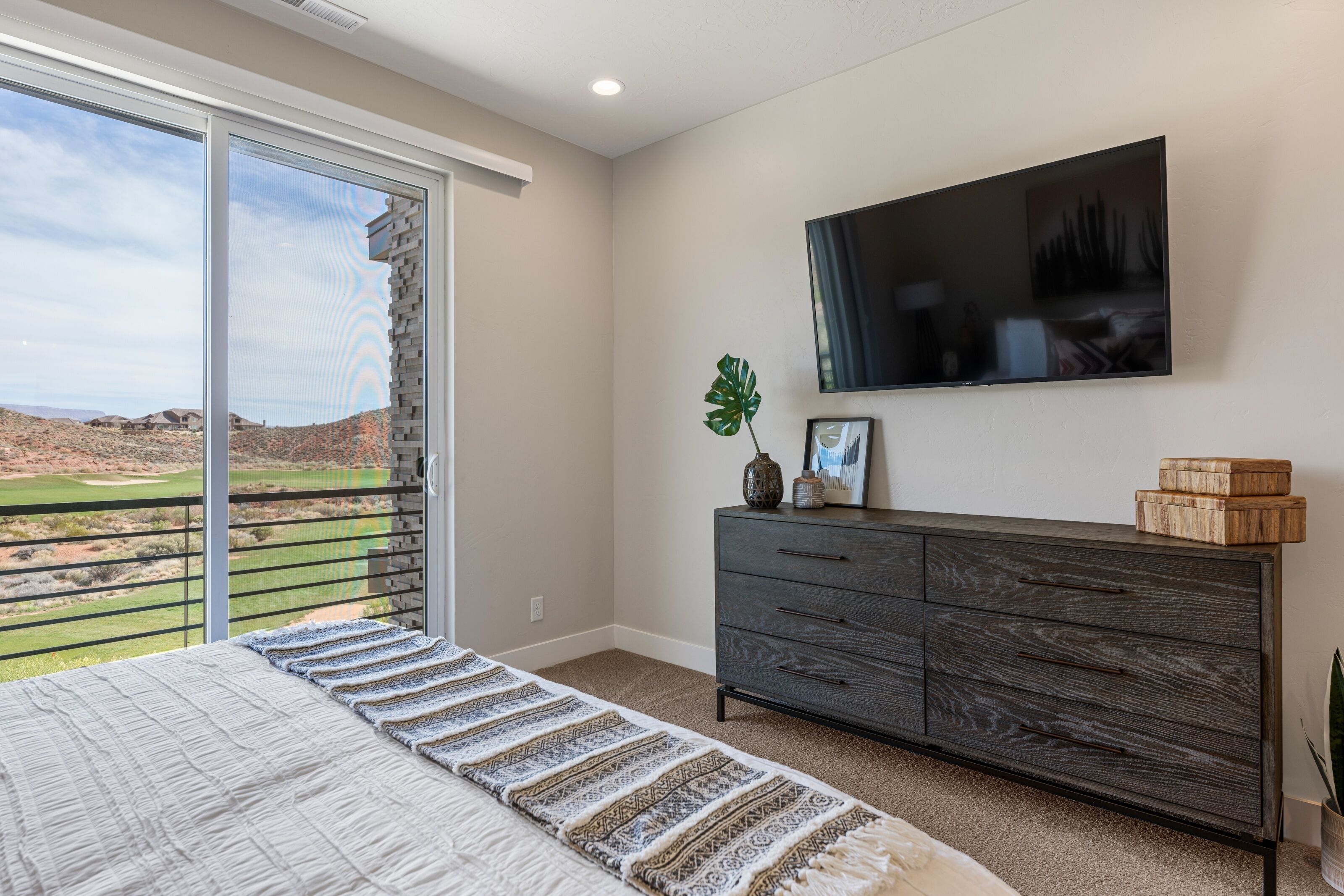 Un dormitorio con una cama grande, un aparador de madera oscura, un televisor de pantalla plana y un ventanal corredero que da a un balcón con vistas a un campo de golf.