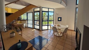 A spacious living area with a wooden staircase, a dining table with chairs, and large sliding glass doors leading to a patio. - Gîte la Majorelle T3 de 92 m² + Terrasses, Jardin (Saint-Guiraud)