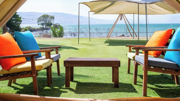 Een overdekte buitenruimte met twee houten stoelen en een tafel, met uitzicht op een strand en bergen.