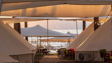 A row of tents with tables and chairs, overlooking a body of water and a mountain.