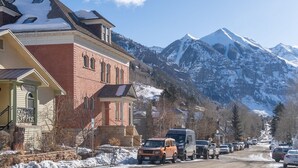 Un edificio con fachada de ladrillo y techo cubierto de nieve, situado en un pueblo montañoso nevado con coches aparcados y montañas al fondo.