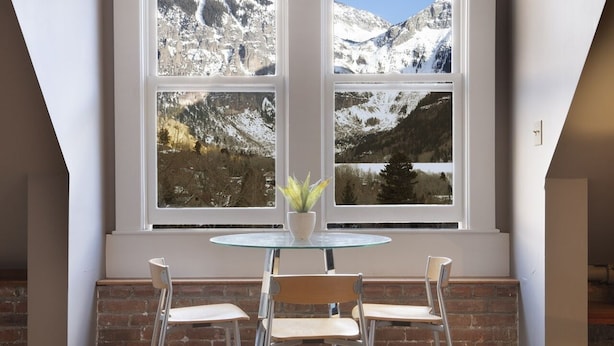 Un comedor con una mesa y sillas, una planta en maceta y vistas a montañas nevadas a través de amplios ventanales.