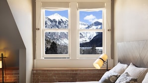 Un dormitorio con un amplio ventanal que ofrece vistas a la montaña, una cama con almohadas estampadas y una lámpara de noche.