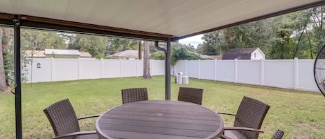 Une terrasse couverte avec une table ronde et des chaises en osier, entourée d’une clôture blanche et d’arbres.