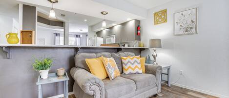 A modern living room with a grey sofa, a small side table with a plant, and a kitchen in the background.
