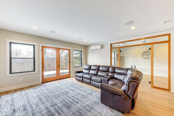A spacious living room with a leather sectional sofa, large windows, and a sliding glass door leading to a balcony. - Mountain-view Home in Stoneham w/ Hot Yoga Studio! (Lovell)