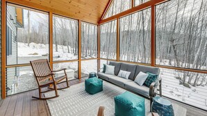 A cozy room with a wooden ceiling, large windows, a gray sofa, teal poufs, and a rocking chair. - Mountain-view Home in Stoneham w/ Hot Yoga Studio! (Lovell)
