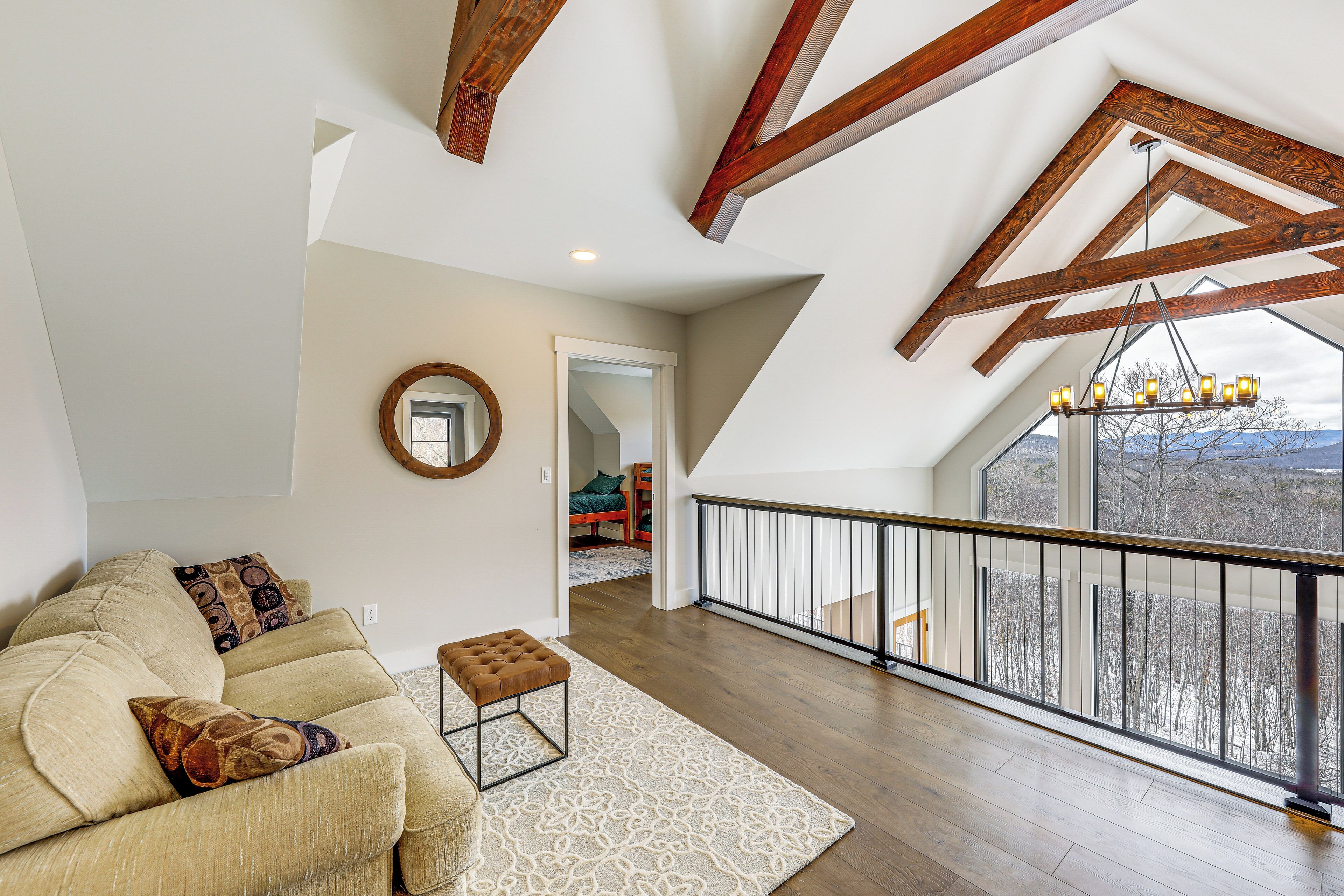 Un loft espacioso con techo alto, vigas de madera a la vista, un amplio ventanal con vistas al exterior, una zona de estar acogedora con sofá y reposapiés, y una escalera que conduce a un nivel superior.