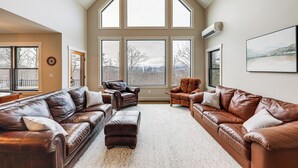 A spacious living room with a high ceiling, large windows, and leather sofas. - Mountain-view Home in Stoneham w/ Hot Yoga Studio! (Lovell)