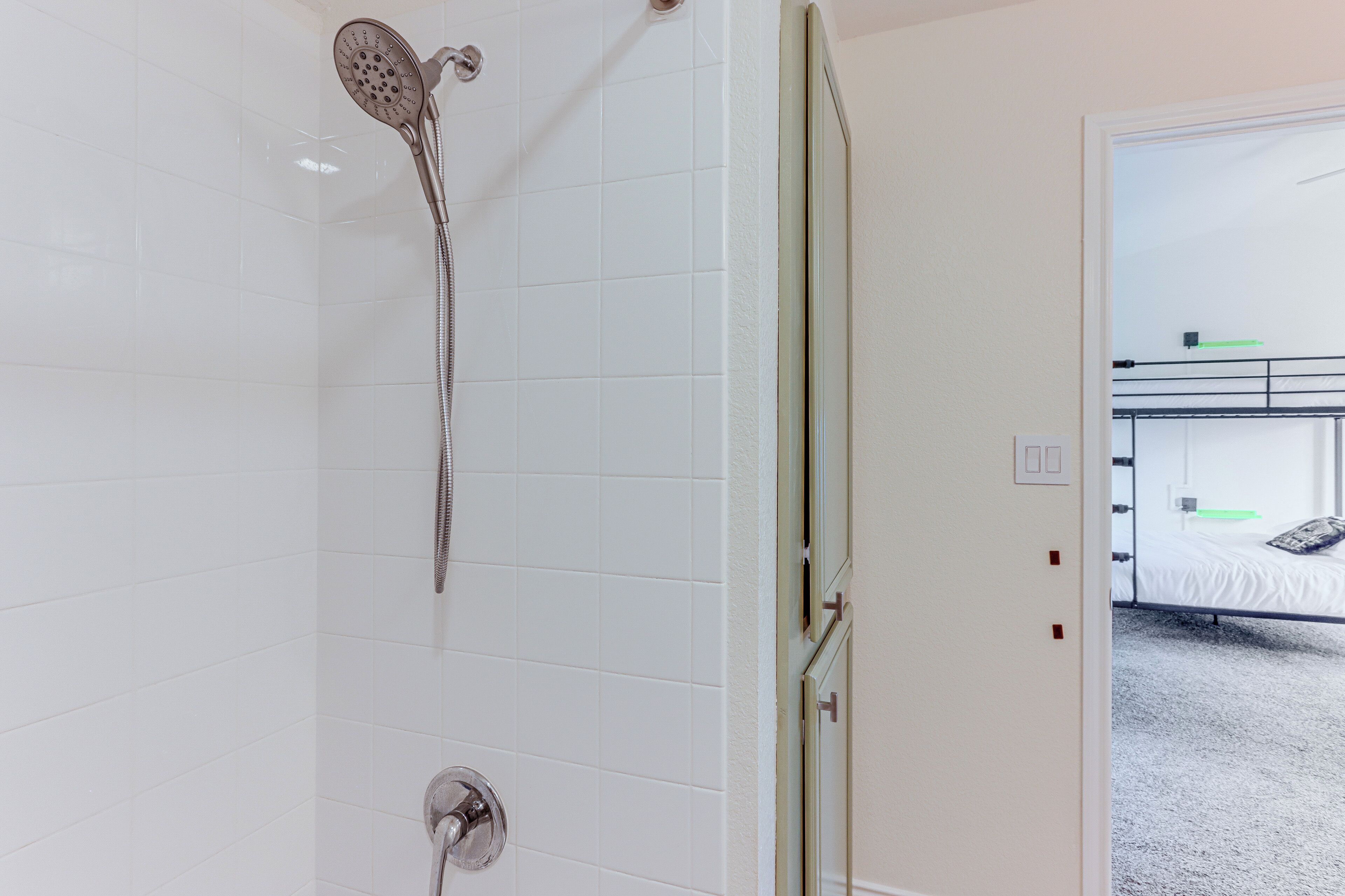 Un baño con azulejos blancos, un cabezal de ducha y una puerta que da a un dormitorio con literas.