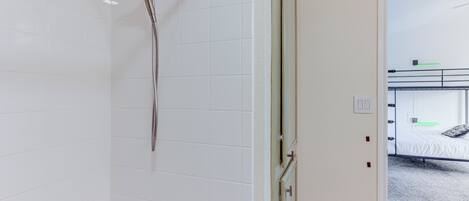 Un baño con azulejos blancos, un cabezal de ducha y una puerta que da a un dormitorio con literas.