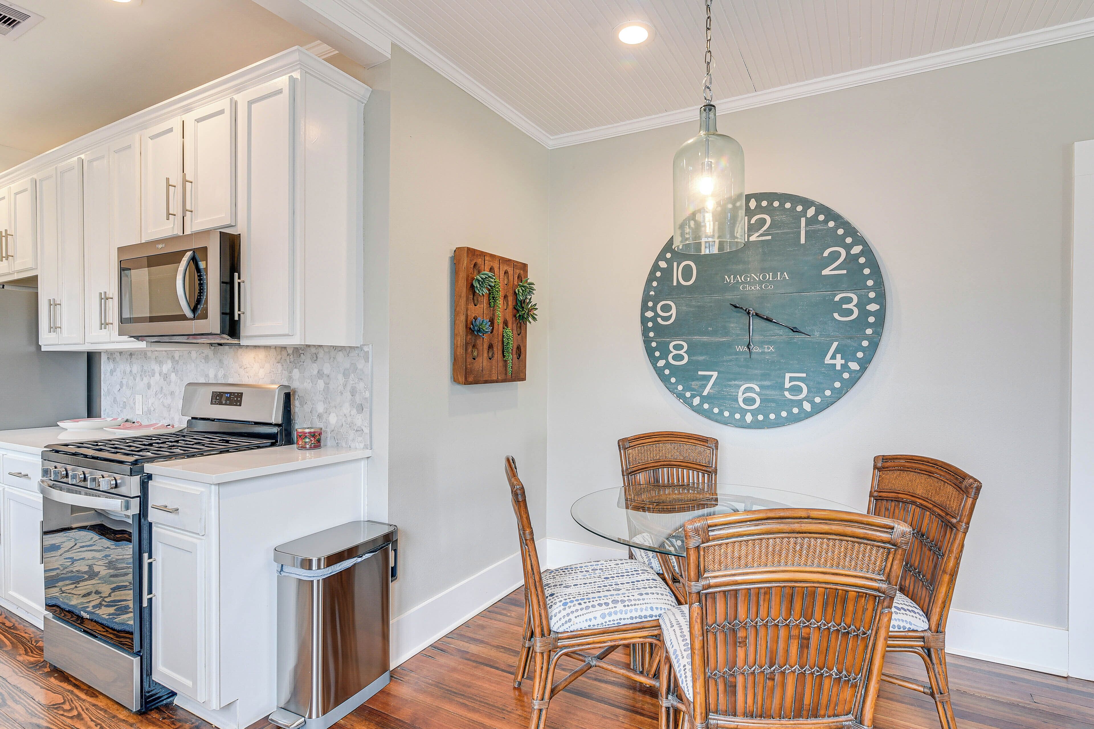 A kitchen with white cabinets, a stainless steel stove, and a microwave. There is a dining table with wicker chairs and a large wall clock.