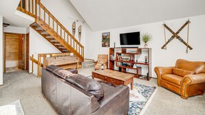 A spacious living room with a wooden staircase, leather sofa, and a television. - Woodland Park Chalet w/ Deck, Grill & Mtn Views! (Woodland Park)