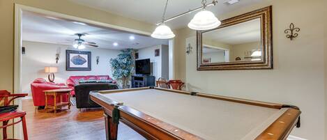 A pool table in a room with wooden flooring, a red chair, a black sofa, and a ceiling fan.