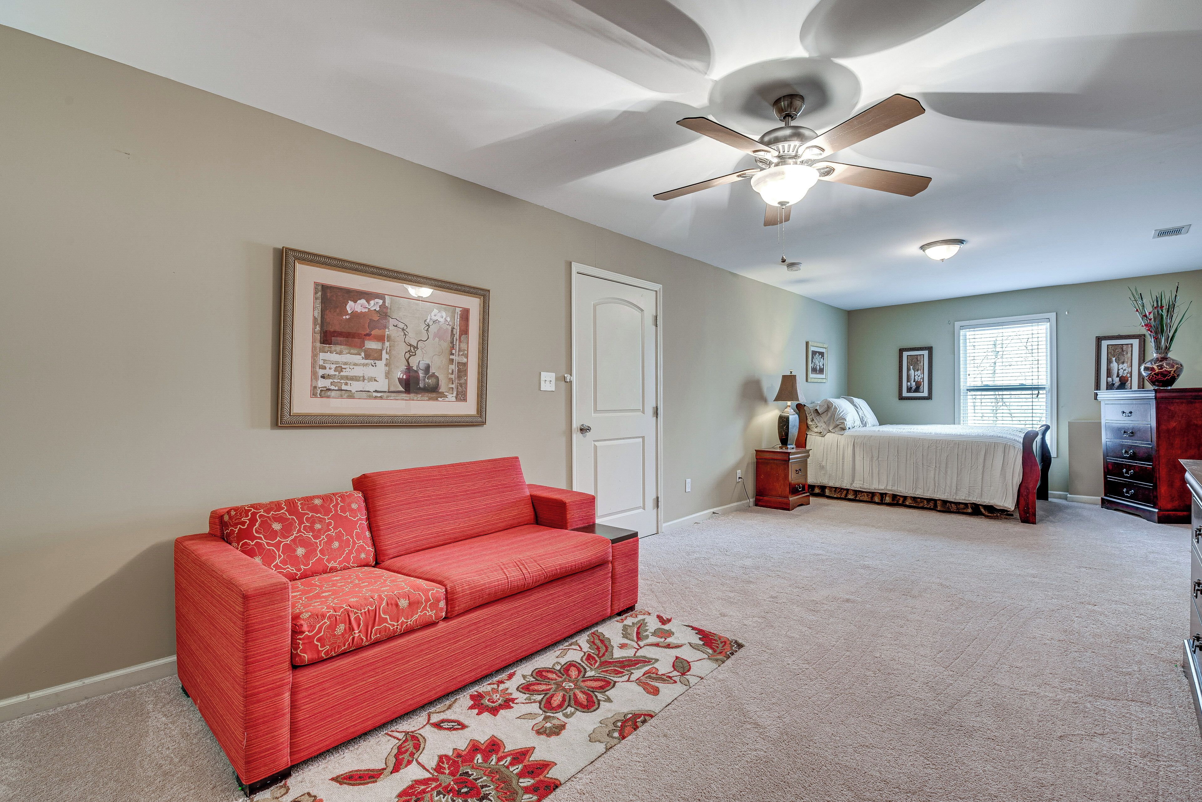 A bedroom with a red sofa, a bed, a ceiling fan, and a window with blinds.