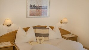 A neatly made bed with white linens, two pillows, and a heart-shaped towel arrangement. There are two bedside lamps and a framed picture on the wall. - Appartment "hochwart" (Nals)