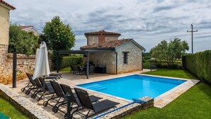 Una piscina con sillas de descanso, zona de barbacoa y edificio de piedra con techo de tejas.