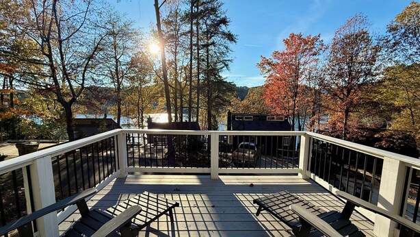 Eine Holzterrasse mit Metallgeländern, zwei Liegestühle und Blick auf Bäume sowie ein Gebäude gegenüber dem Wasser.