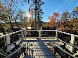 A wooden deck with metal railings, two lounge chairs, and a view of trees and a building across a body of water.