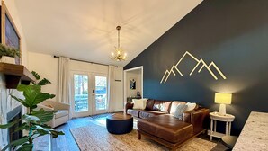 Ein modernes Wohnzimmer mit Kamin, einer großen Pflanze, einem Ledersofa, einem Couchtisch und einem Teppich. Durch die Glastüren bietet sich ein Blick ins Freie.