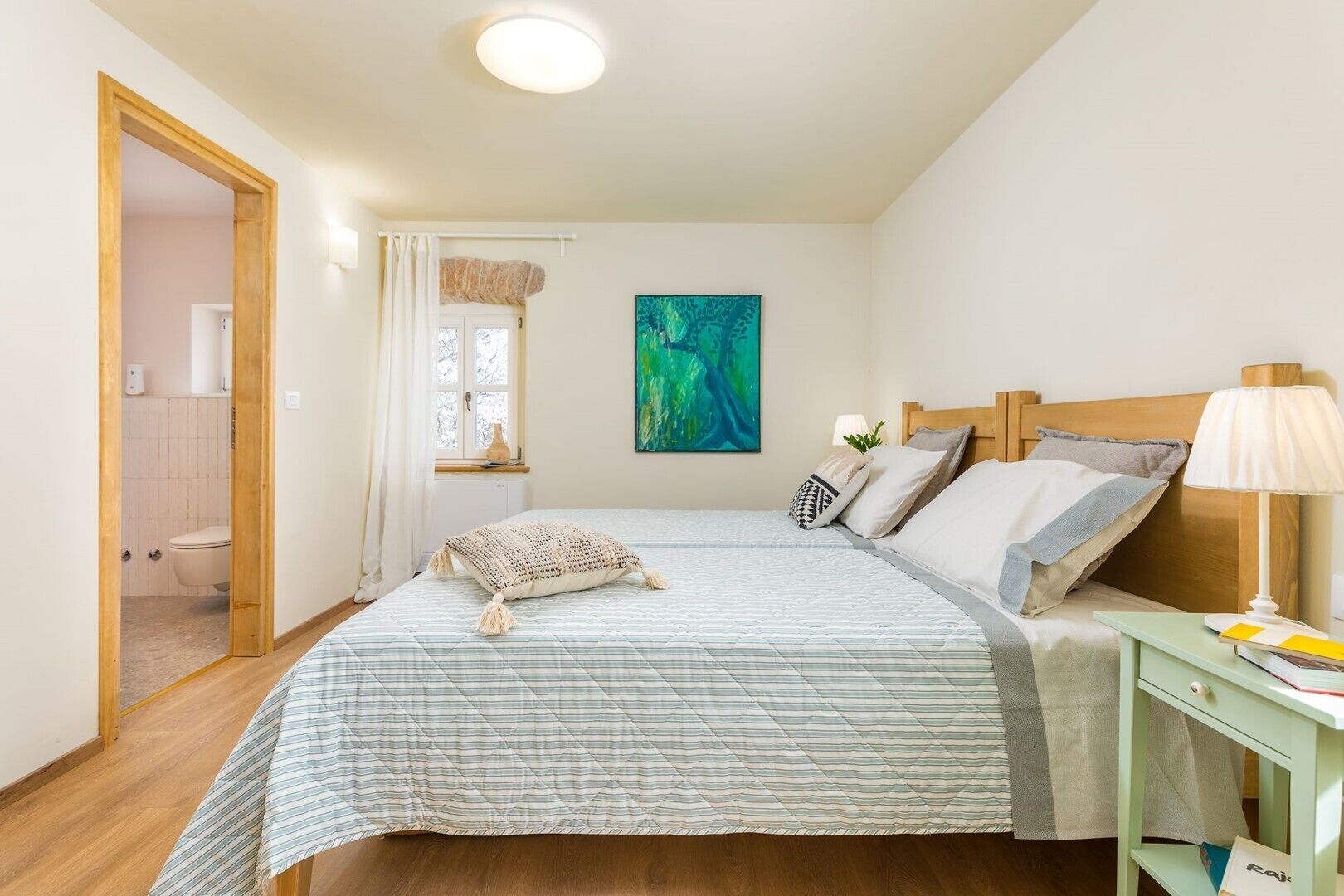 A bedroom with a bed, a nightstand, a lamp, a painting, and a view of a bathroom.