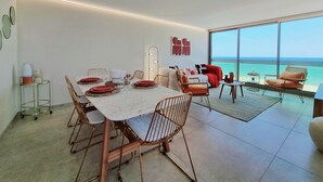 Una zona de menjador moderna amb un gran taulell preparat per a sis persones, envoltat de cadires metàl·liques. Un sofà i un petit taulell rodó es veuen al fons, amb un gran finestral que ofereix vista a l'oceà.