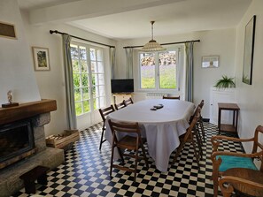 A dining room with a checkered floor, a fireplace, a television, and a view of the garden through the windows. - Maison de Vacances à Louer sur L'ile Callot - Ker Douara (Carantec)